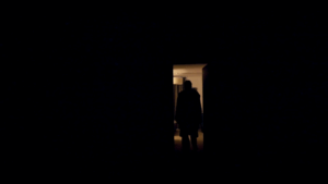 Moody silhouette of a person standing in a doorway leading into a dimly lit room