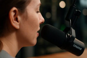Close-up, cinematic view over a woman’s shoulder showing her face in soft focus as she speaks into a Shure-style podcast microphone, with blurred studio lights and cameras in the background — symbolising professional podcast production and authentic storytelling.